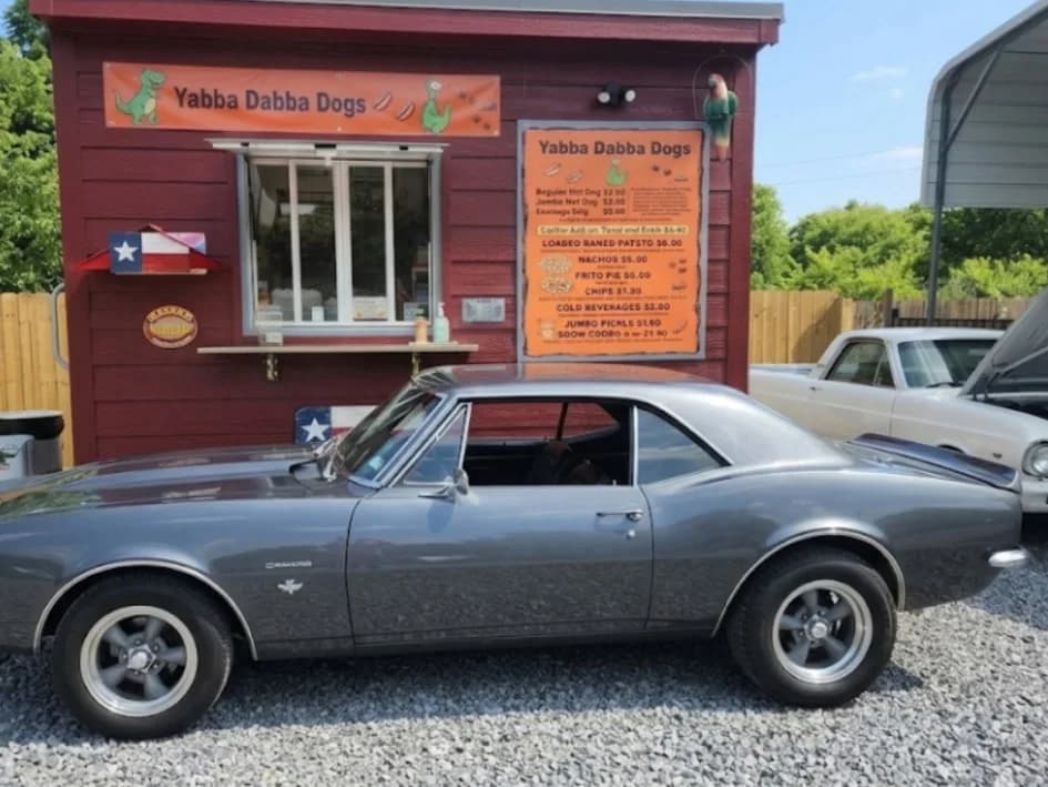 A gray 1967 Chevrolet Camaro parked in front of the Yabba Dabba Dogs food truck and its menu board in Nevada, Texas