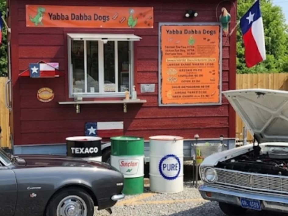 The Yabba Dabba Dogs food truck storefront with a Texas flag, vintage Texaco, Sinclair, and Pure oil drums, and classic cars parked out front
