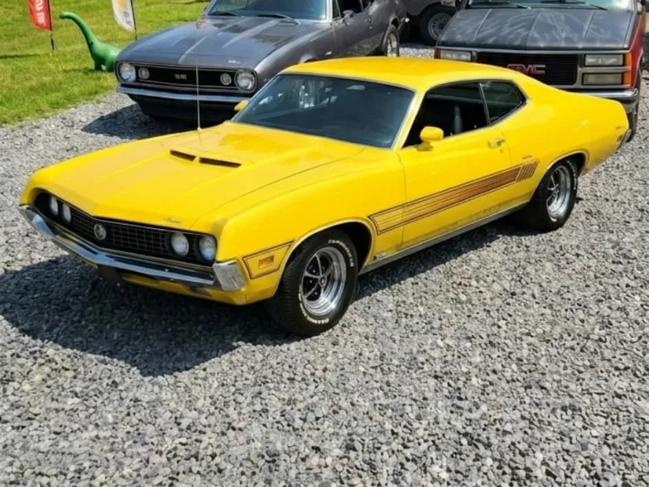 A bright yellow 1970 Ford Torino GT with other classic muscle cars on the gravel lot at Yabba Dabba Dogs